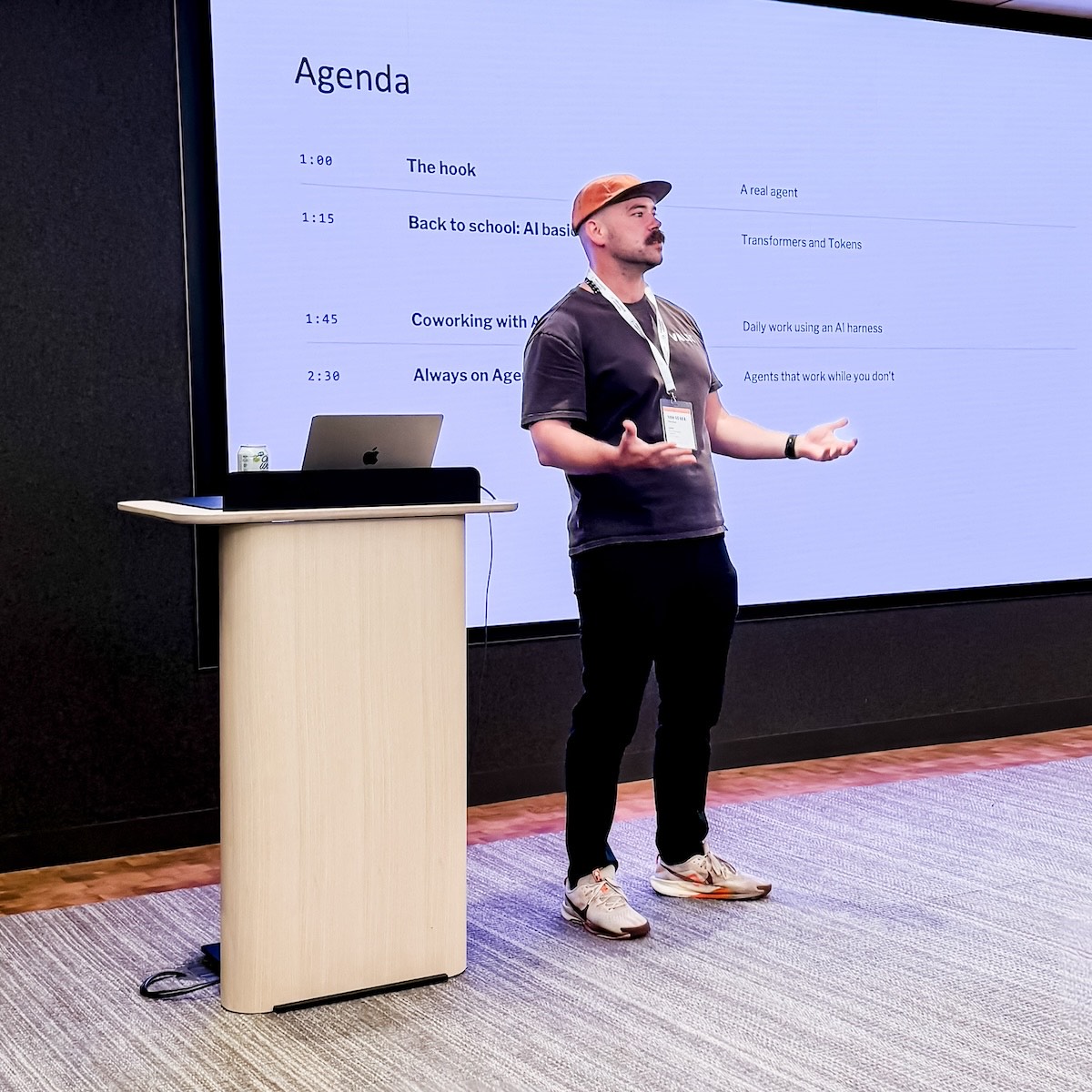 Michael Ducker on stage at an AI training session, talking through the agenda slide behind him: the hook, AI basics, coworking with AI, and always-on agents